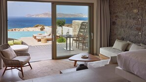 A modern room with a large sliding glass door leading to a balcony with a view of the sea and a pool. There is a sofa, a chair, and a small table with a vase. -  Hotel Milos Sea Resort (Milos)