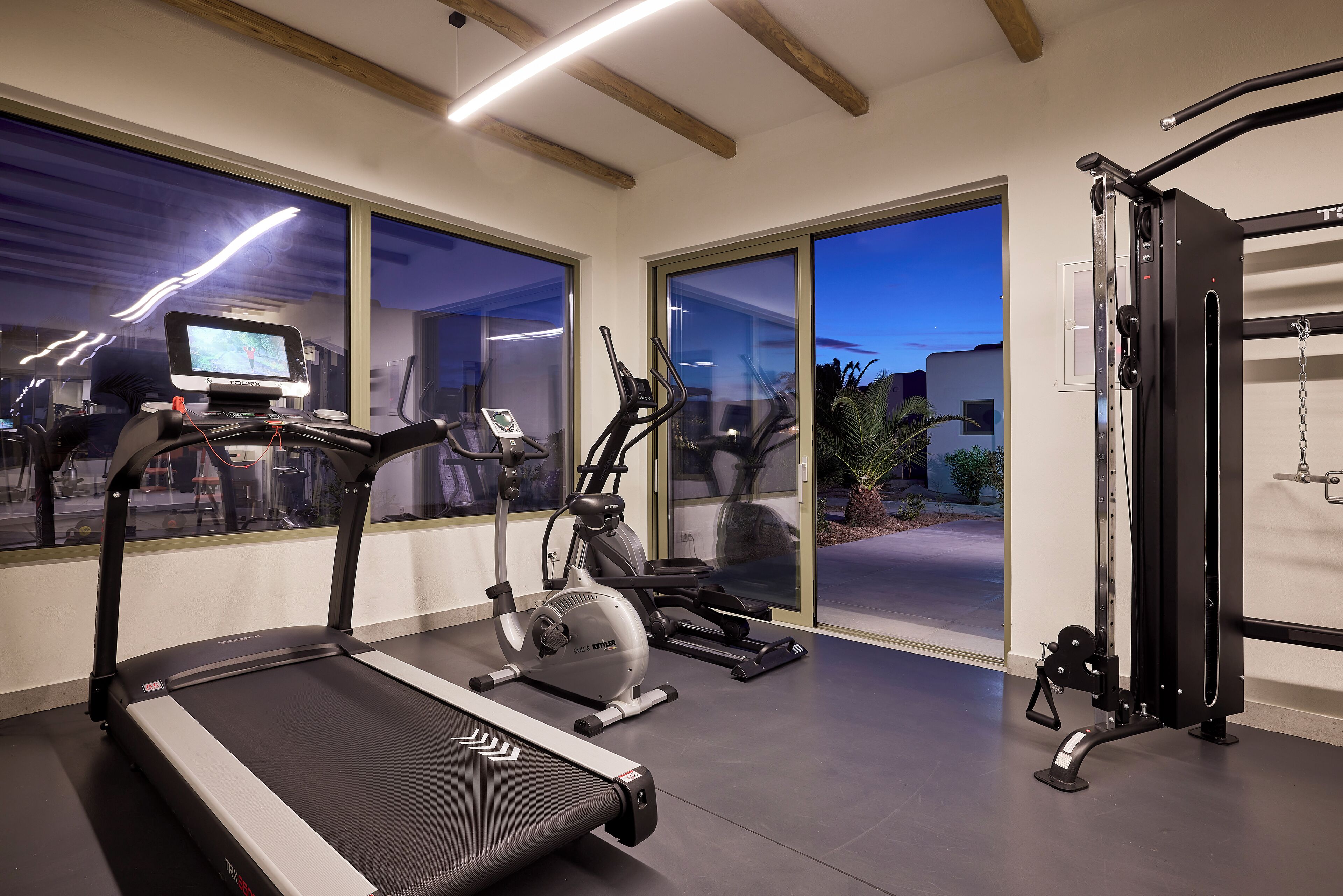 Ein Fitnessstudio mit Laufbändern, Crosstrainern und einem Krafttrainer, ausgestattet mit großen Fenstern und moderner Deckenkonstruktion.