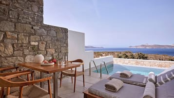 Eine Steinsichtmauer, ein Holztisch mit Stühlen, ein Pool mit Liegestühlen und Meerblick.