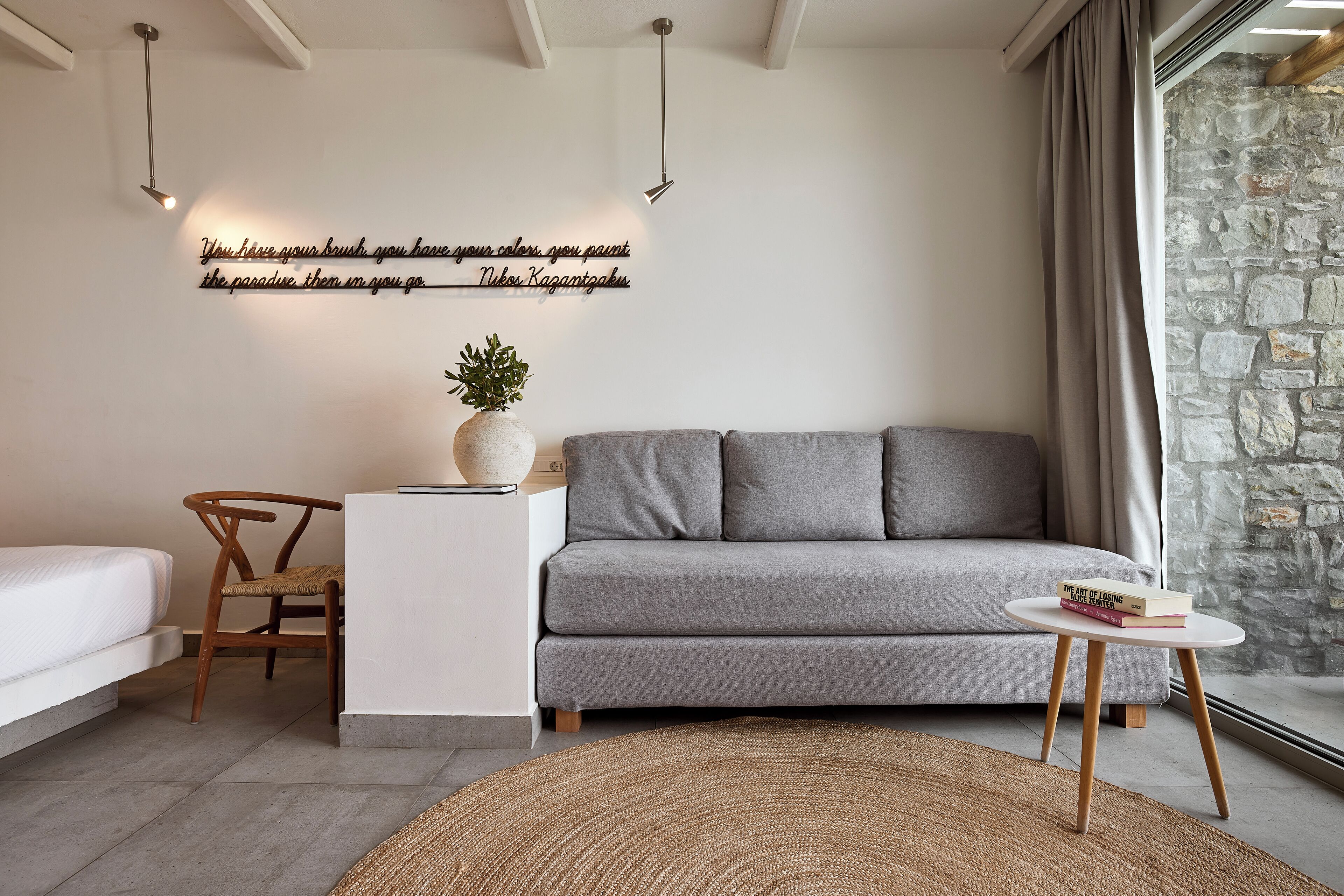 A modern living room with a grey sofa, a small white side table, a wicker chair, a round rug, and a wall with a motivational quote.