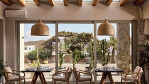 A dining area with large windows, wicker pendant lights, and a view of cacti and buildings outside. -  Hotel Milos Sea Resort (Milos)