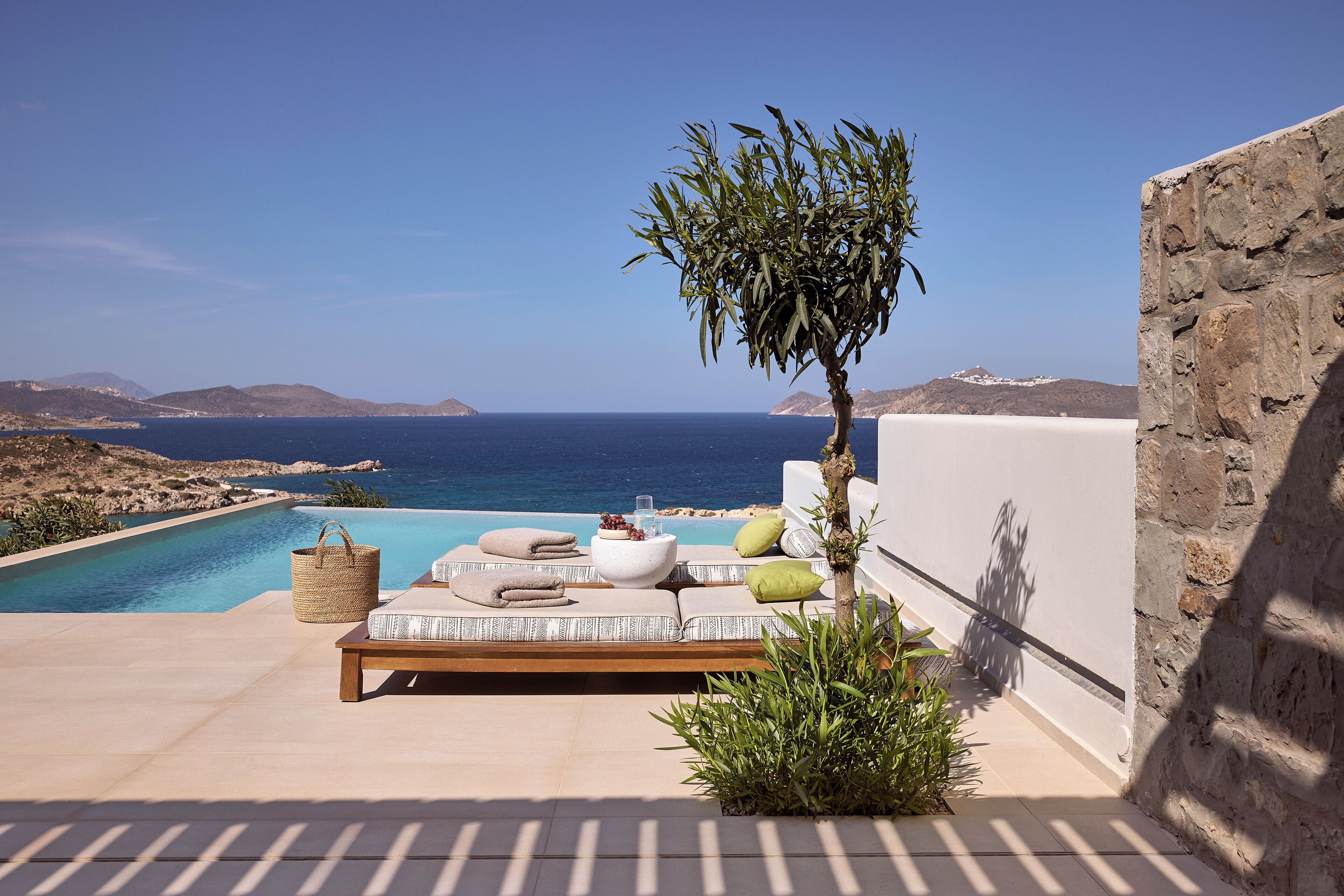 A sunlit outdoor seating area with a pool, stone wall, and a view of the sea.