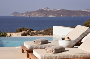 Un área de descanso junto a la piscina con dos sillones acolchados, una mesa redonda con un vaso y fruta, y vistas al mar y montañas a lo lejos.