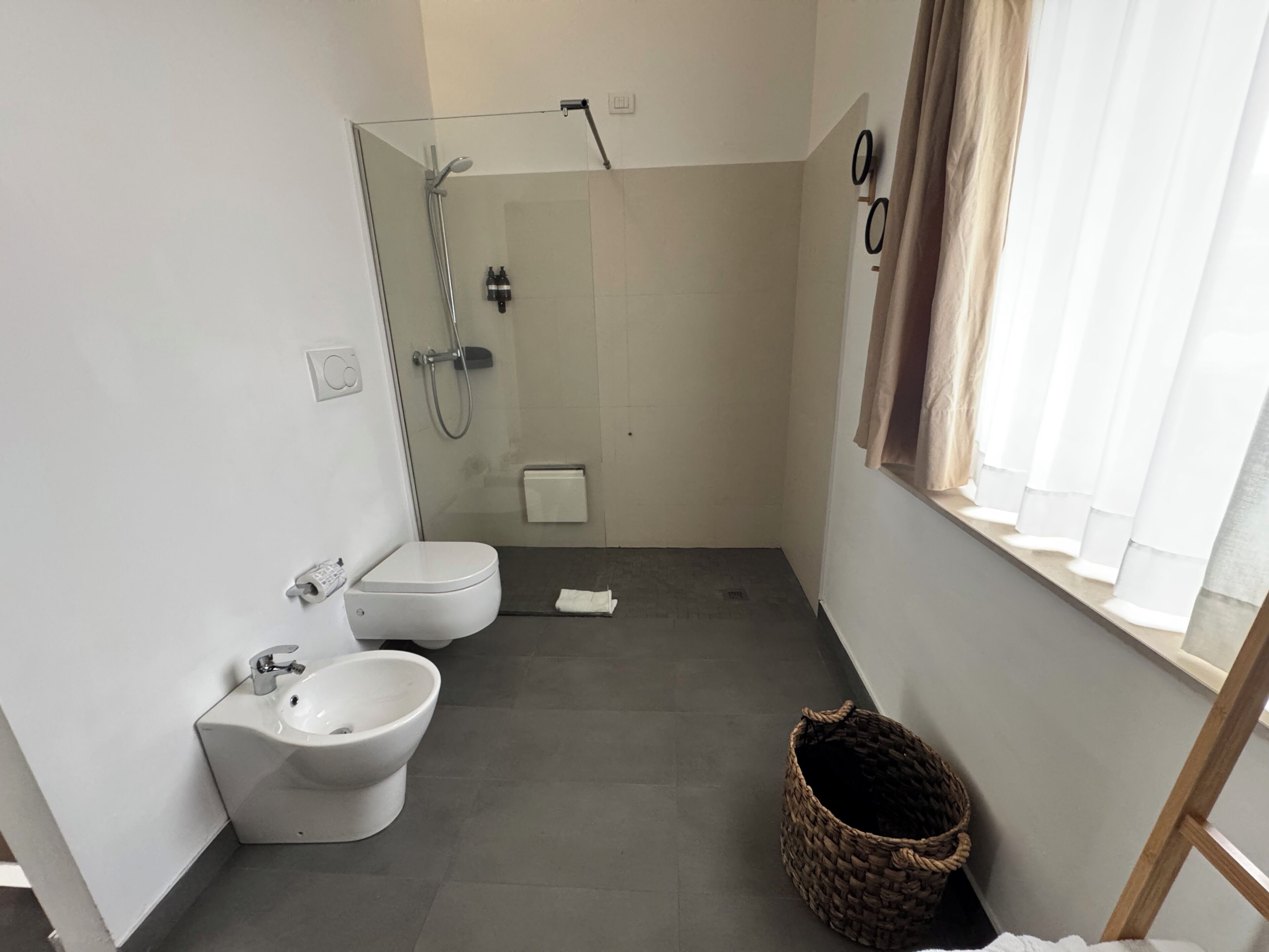 Un baño moderno con ducha, inodoro, bidé y una pequeña ventana con cortinas.