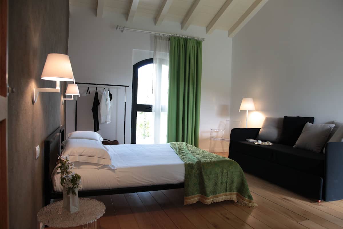 Un dormitorio con cama, lámparas de noche, una cortina verde, un sofá negro y una pequeña mesa redonda con un jarrón.