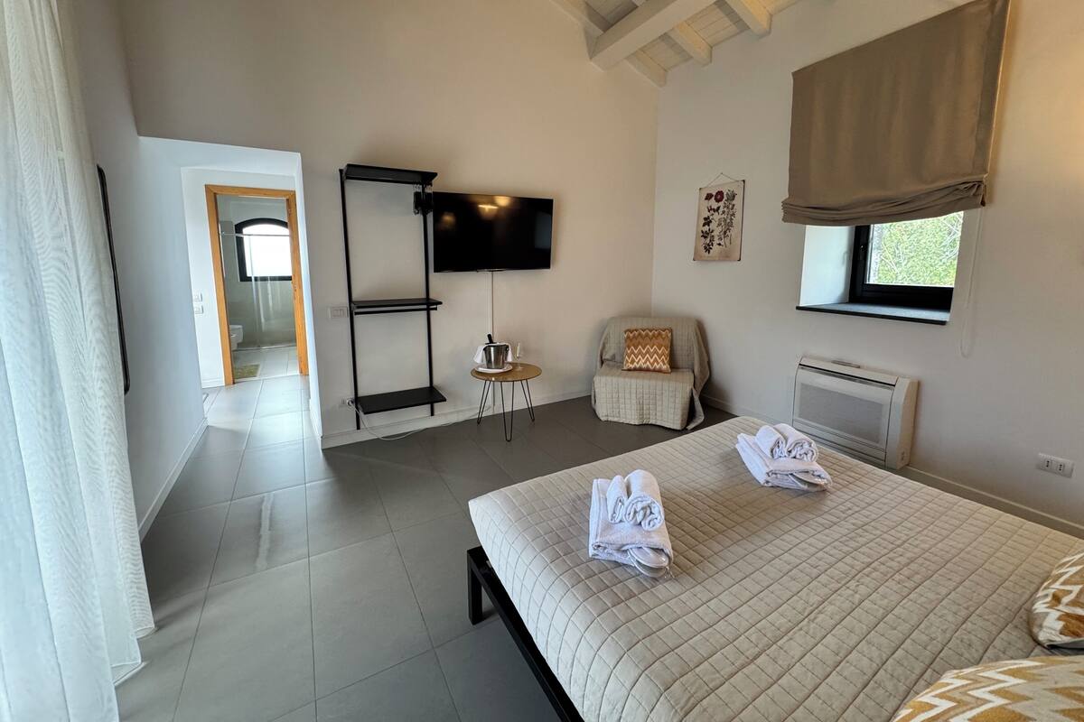 Un dormitorio con cama, televisor, silla, ventana y radiador.