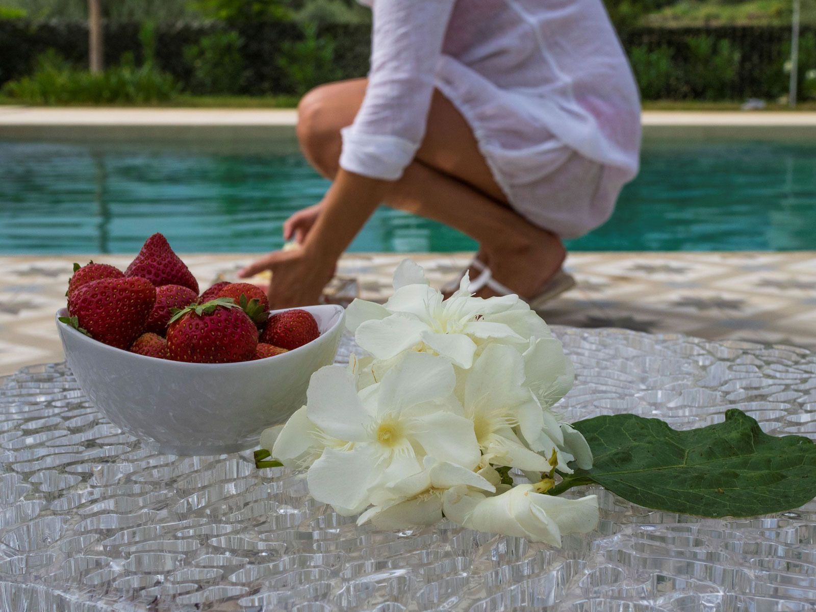 A bowl of strawberries and white flowers on a glass table by a pool.