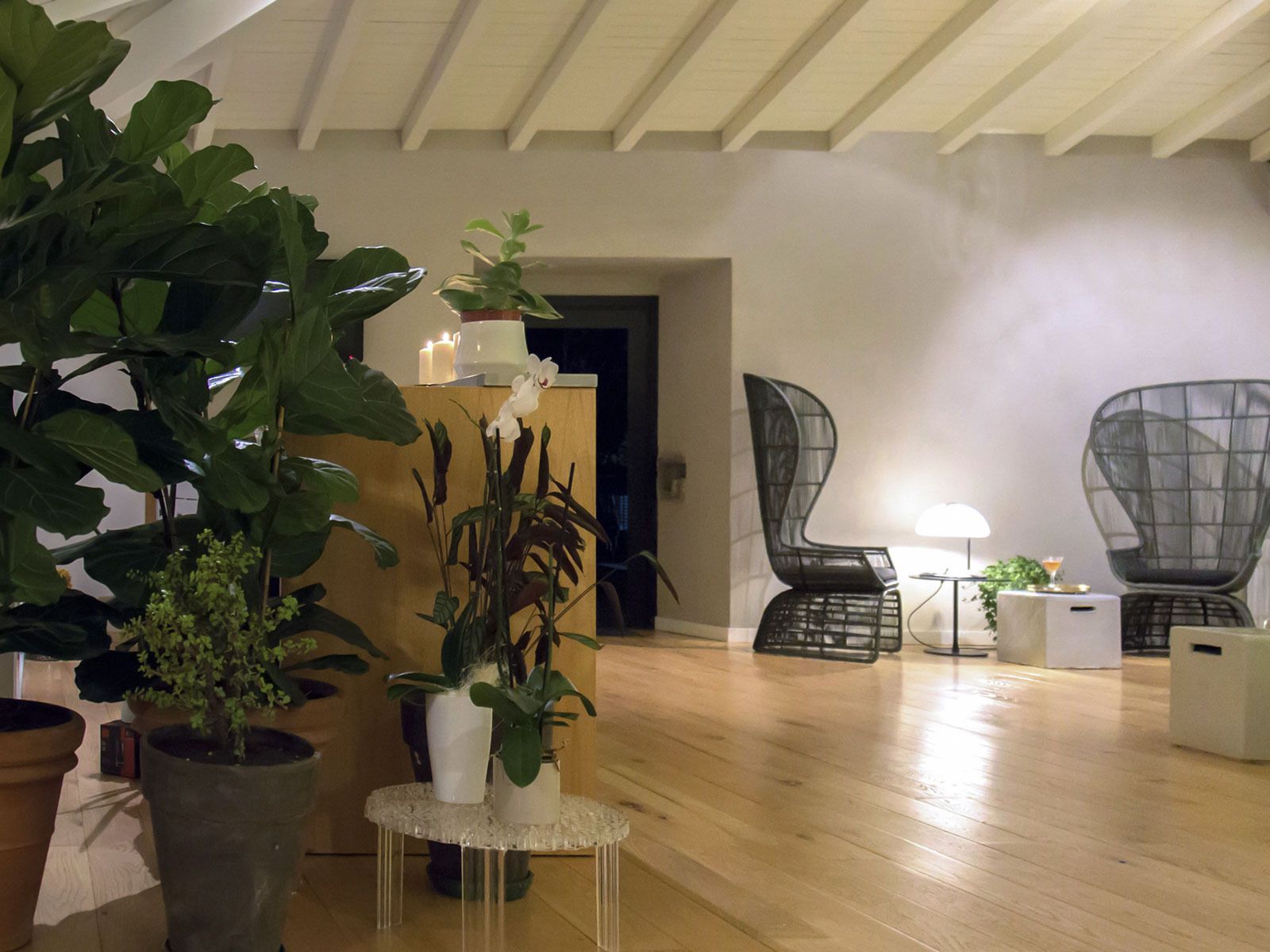 Un salon moderne avec un plancher en bois, une table ronde en verre et deux chaises. On y trouve des plantes en pot et une bougie allumée sur une étagère.