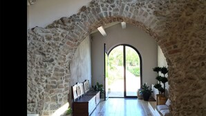 Un arco de piedra conduce a una habitación luminosa con una puerta de cristal, plantas en macetas y un banco de madera.