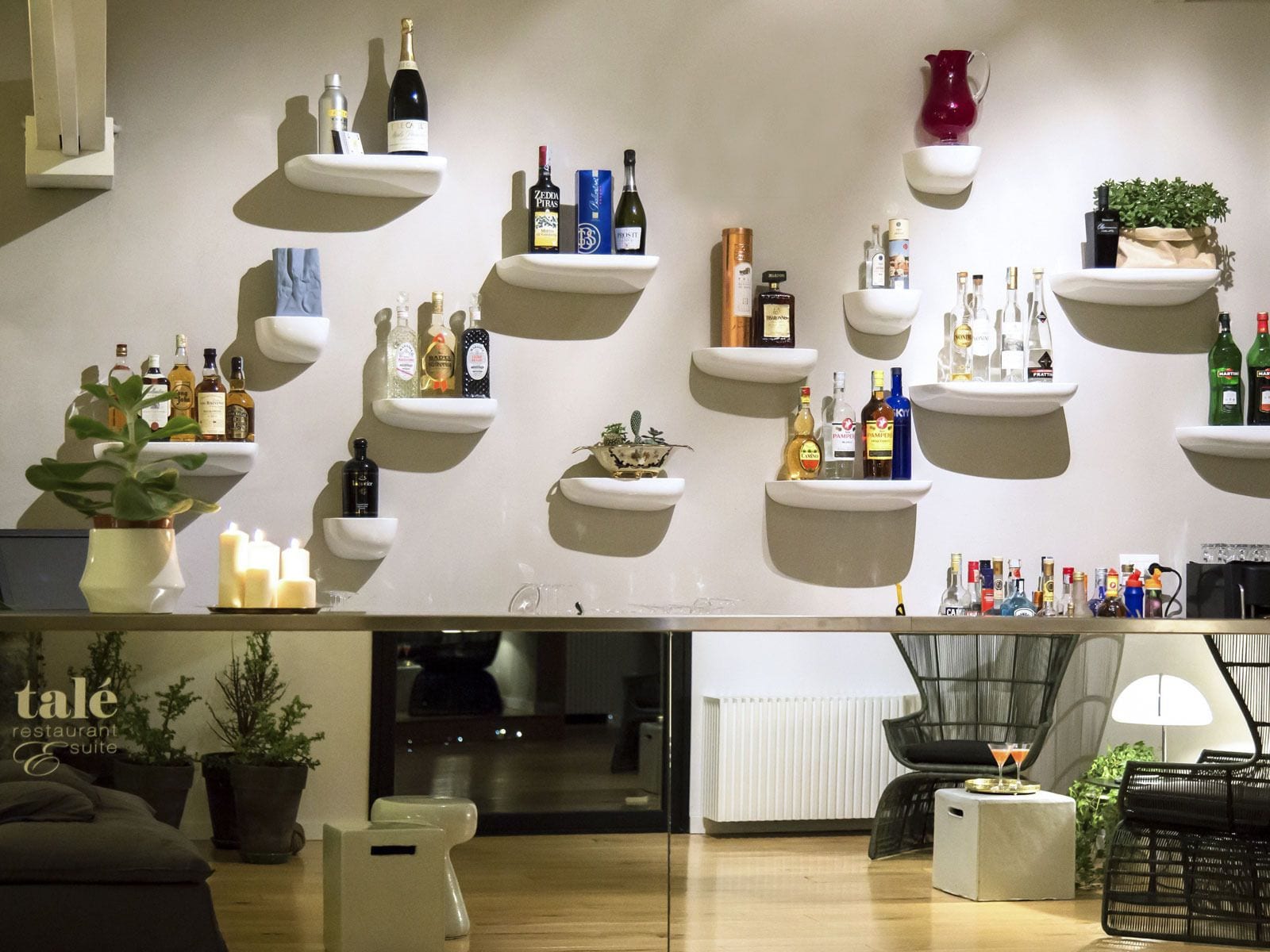 Un espace bar moderne avec des étagères remplies de différentes bouteilles, un coin salon avec un canapé et des fauteuils, et une plante en pot.