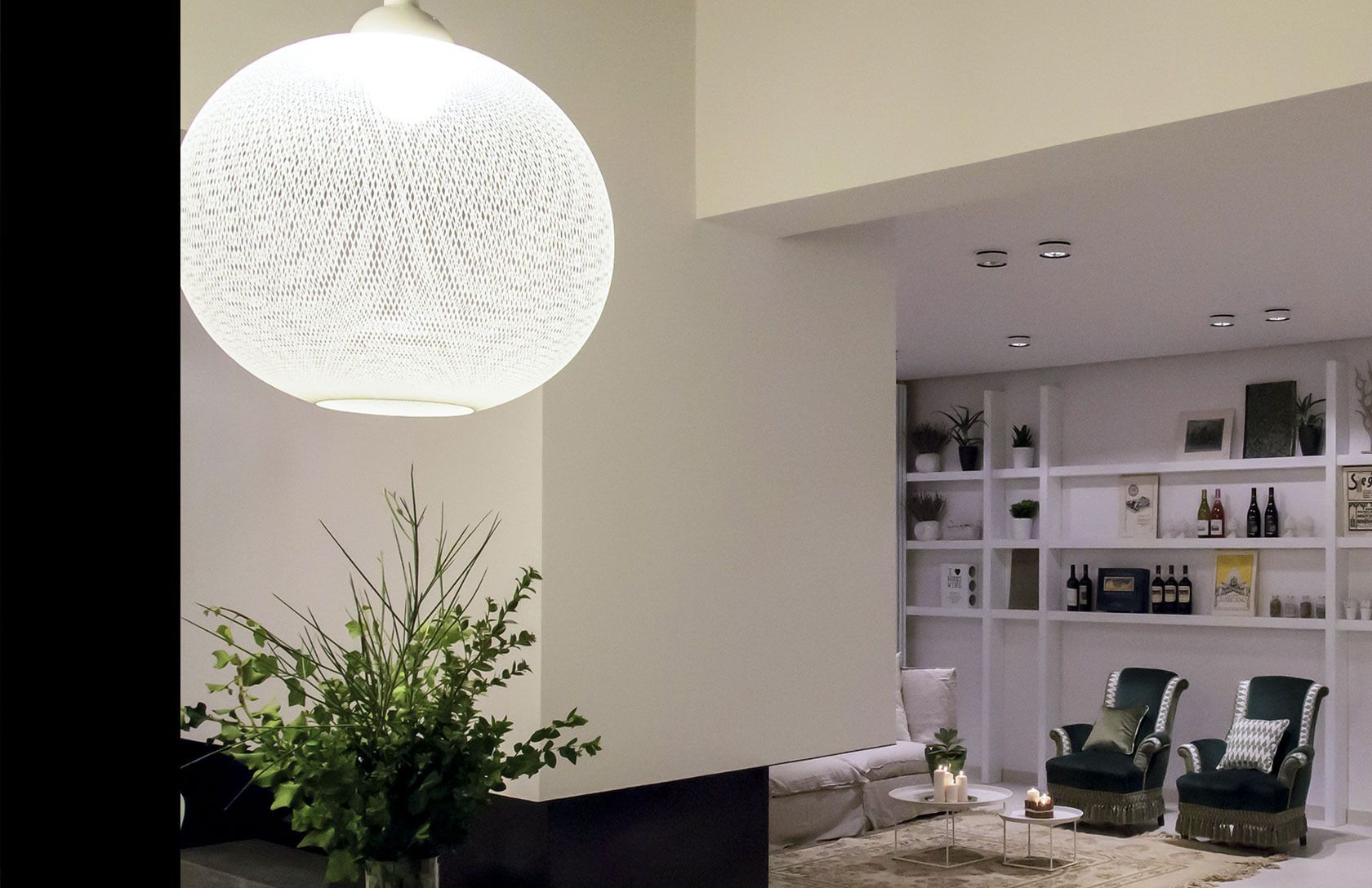 Un salon moderne avec un luminaire rond à texture intéressante, une plante verte, un canapé, deux fauteuils, une table basse et une bibliothèque contenant divers objets.