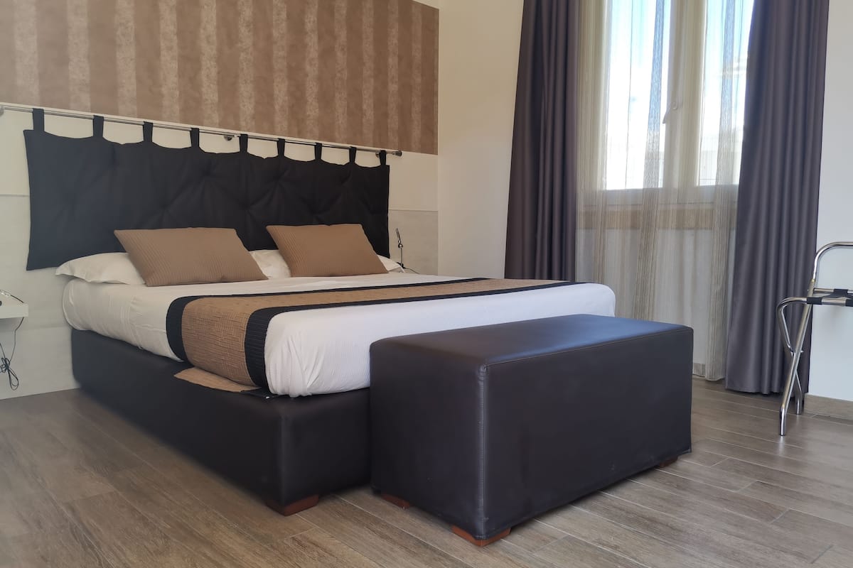 A modern hotel room with a large bed, a black ottoman, and a view of the city through sheer curtains.