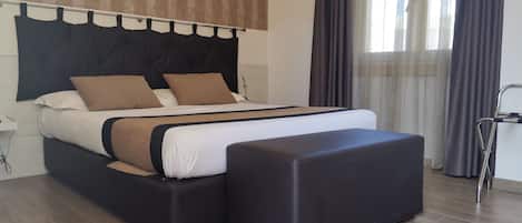 Een moderne hotelkamer met een groot bed, een zwart poef en uitzicht op de stad door dunne gordijnen.