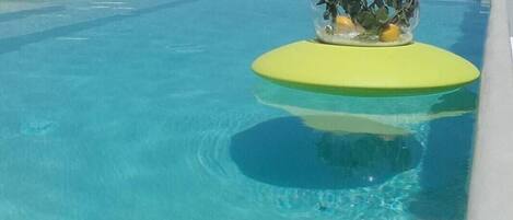Una piscina azul cristalina con una esfera de vidrio sobre un flotador amarillo, rodeada por una zona junto a la piscina en blanco y vistas al mar y a las montañas al fondo.