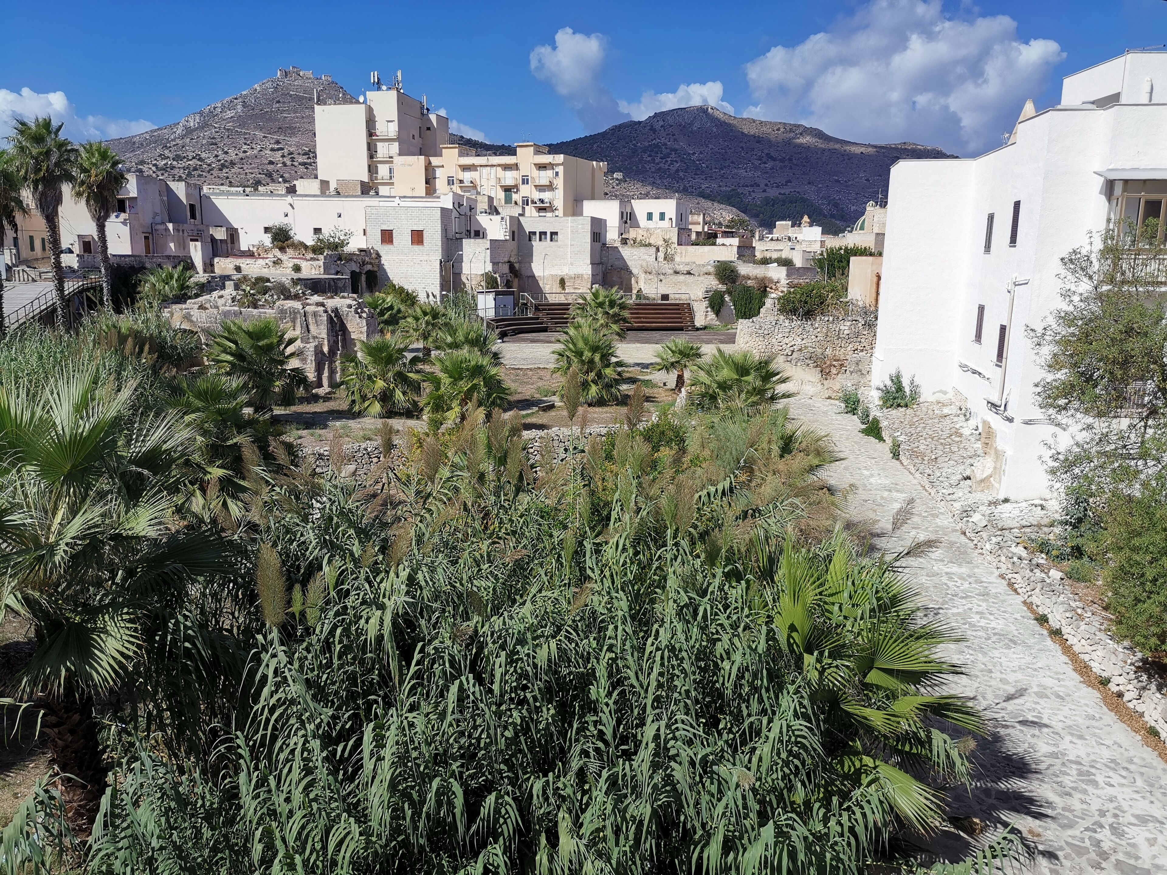 A coastal town with white buildings, a mountain in the background, and a pathway lined with greenery.