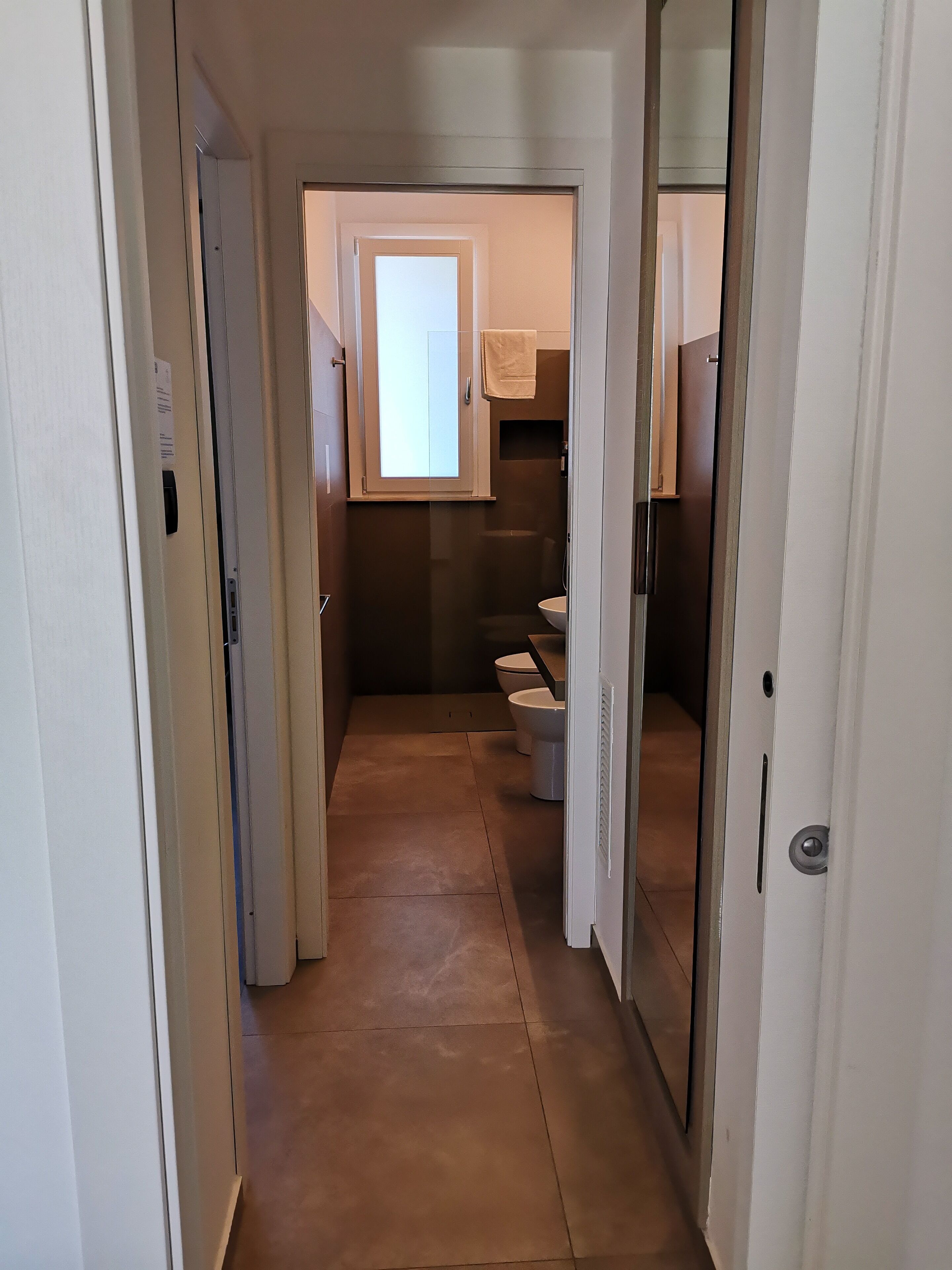Un couloir étroit mène à une salle de bain équipée de toilettes, d’une douche et d’une petite fenêtre.