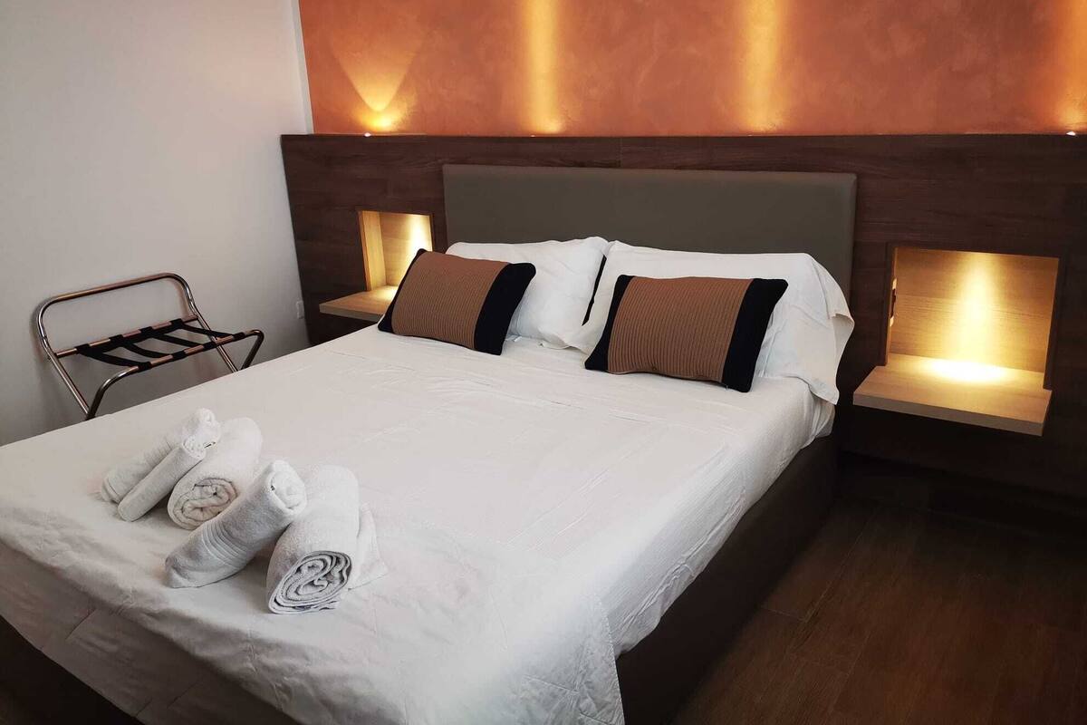 A neatly made bed with white linens and two brown pillows, a metal chair with a towel rack, and two bedside tables with wall-mounted lamps.