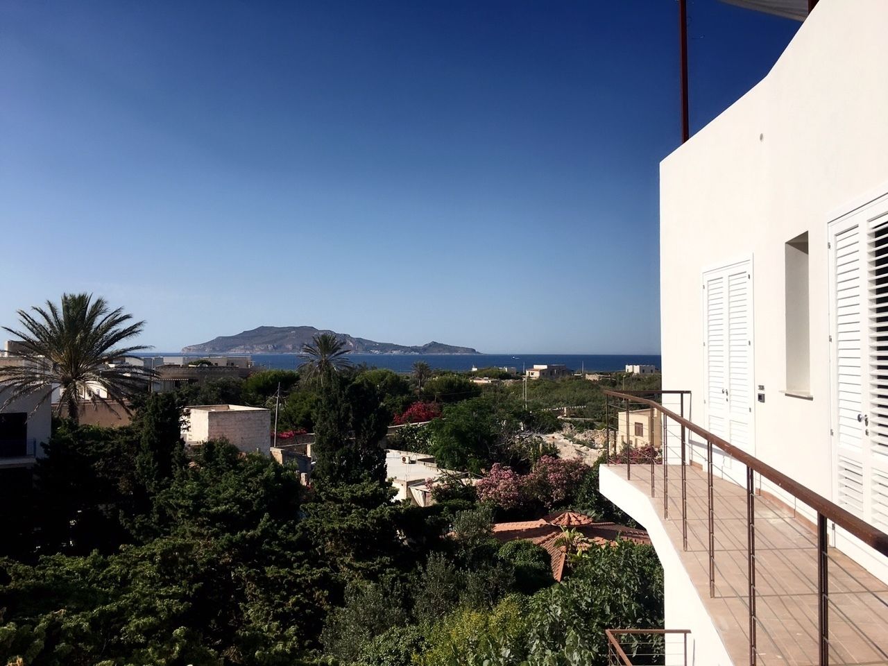 A residential area with a view of the sea, a mountain in the distance, and a white building with shutters.
