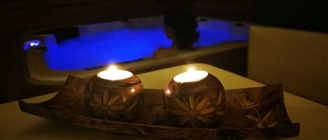 A dimly lit room with a wooden table featuring two candles. In the background, there is a blue-lit bathtub.