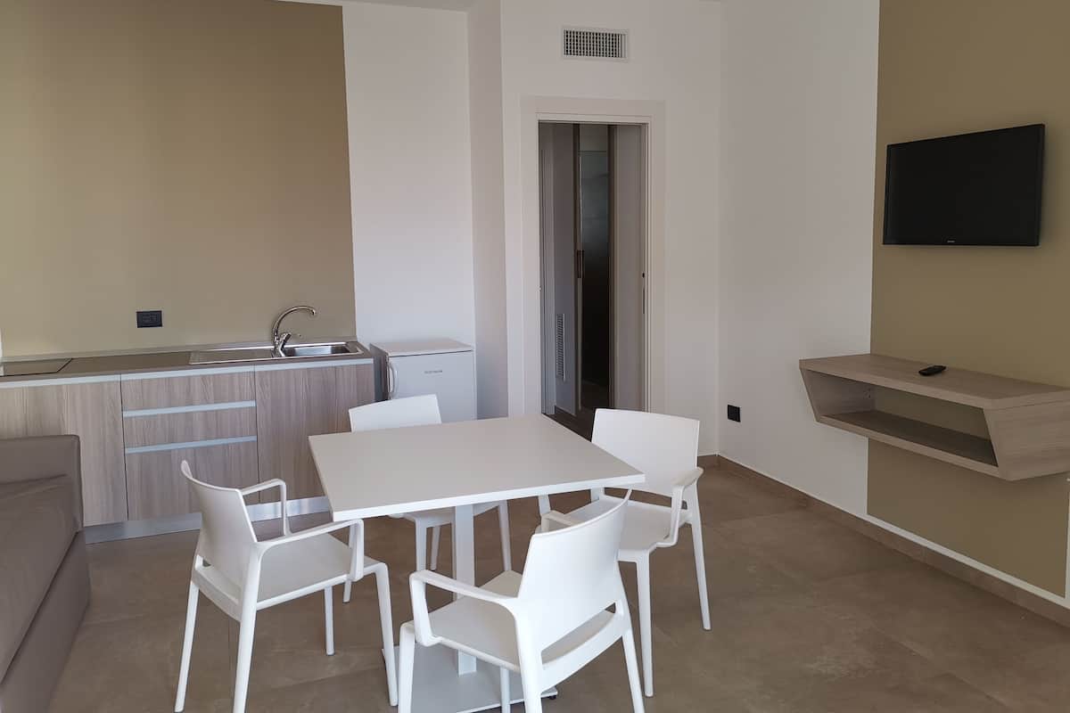 A modern kitchen with a built-in sink and a compact refrigerator. There is a dining area with a white table and chairs. A flat-screen TV is mounted on the wall above a wooden shelf.
