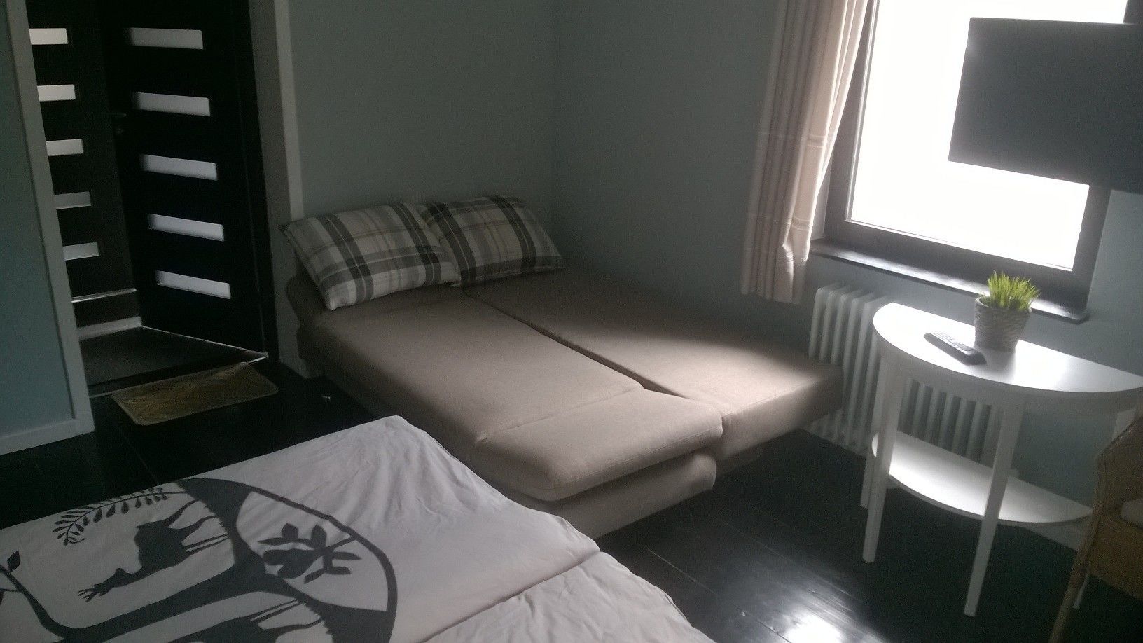 Een kleine kamer met een bed, een tafeltje met een plant en een radiator.