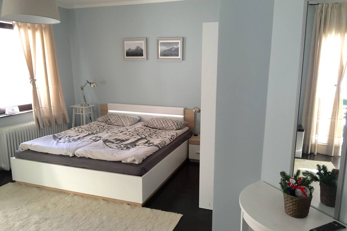 A bedroom with a bed, a bedside table, a lamp, a nightstand, a window with curtains, and a small table with a remote control and a decorative item.