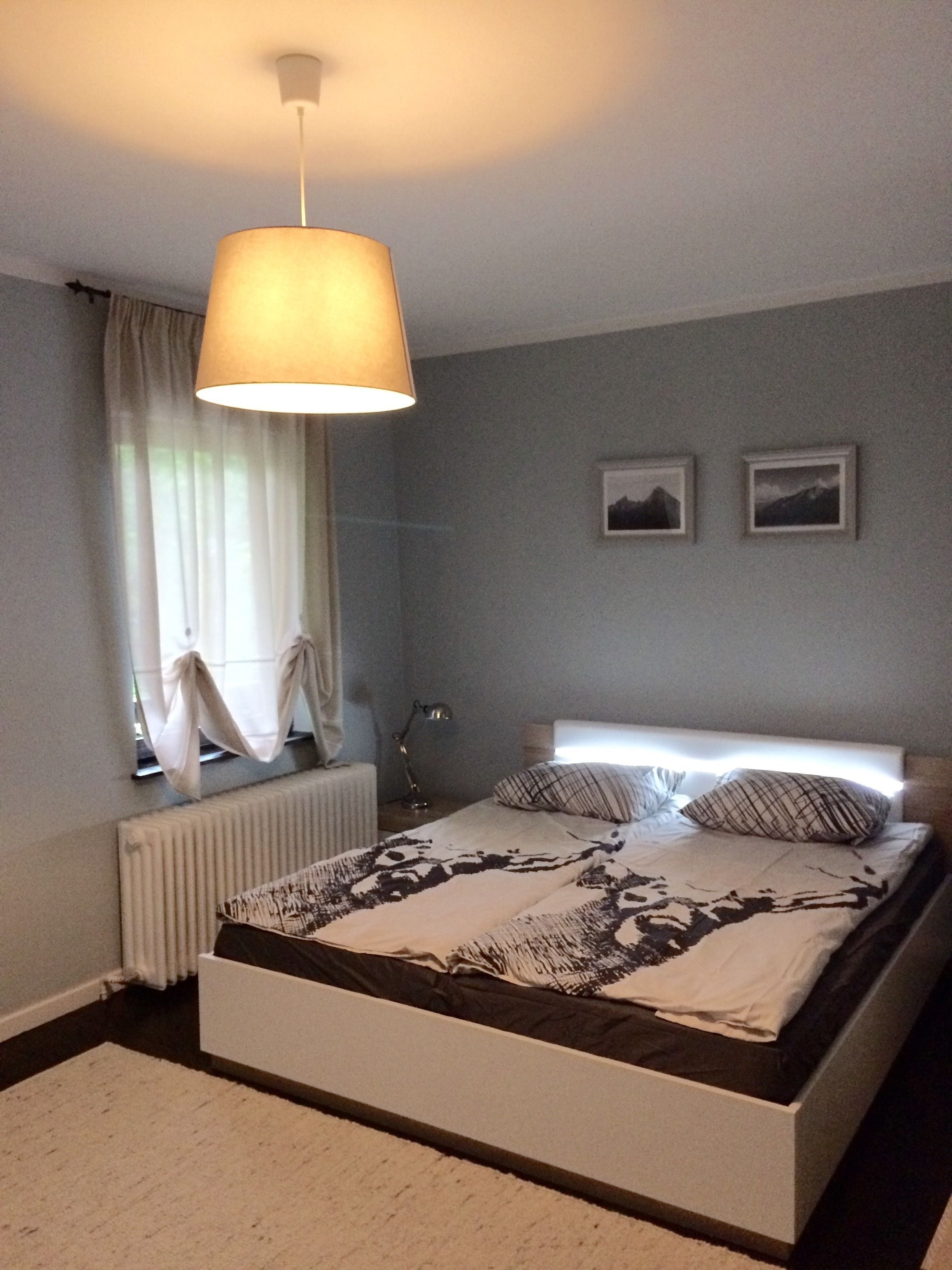 Un dormitorio con cama, ventana con cortinas, radiador y dos cuadros en la pared.