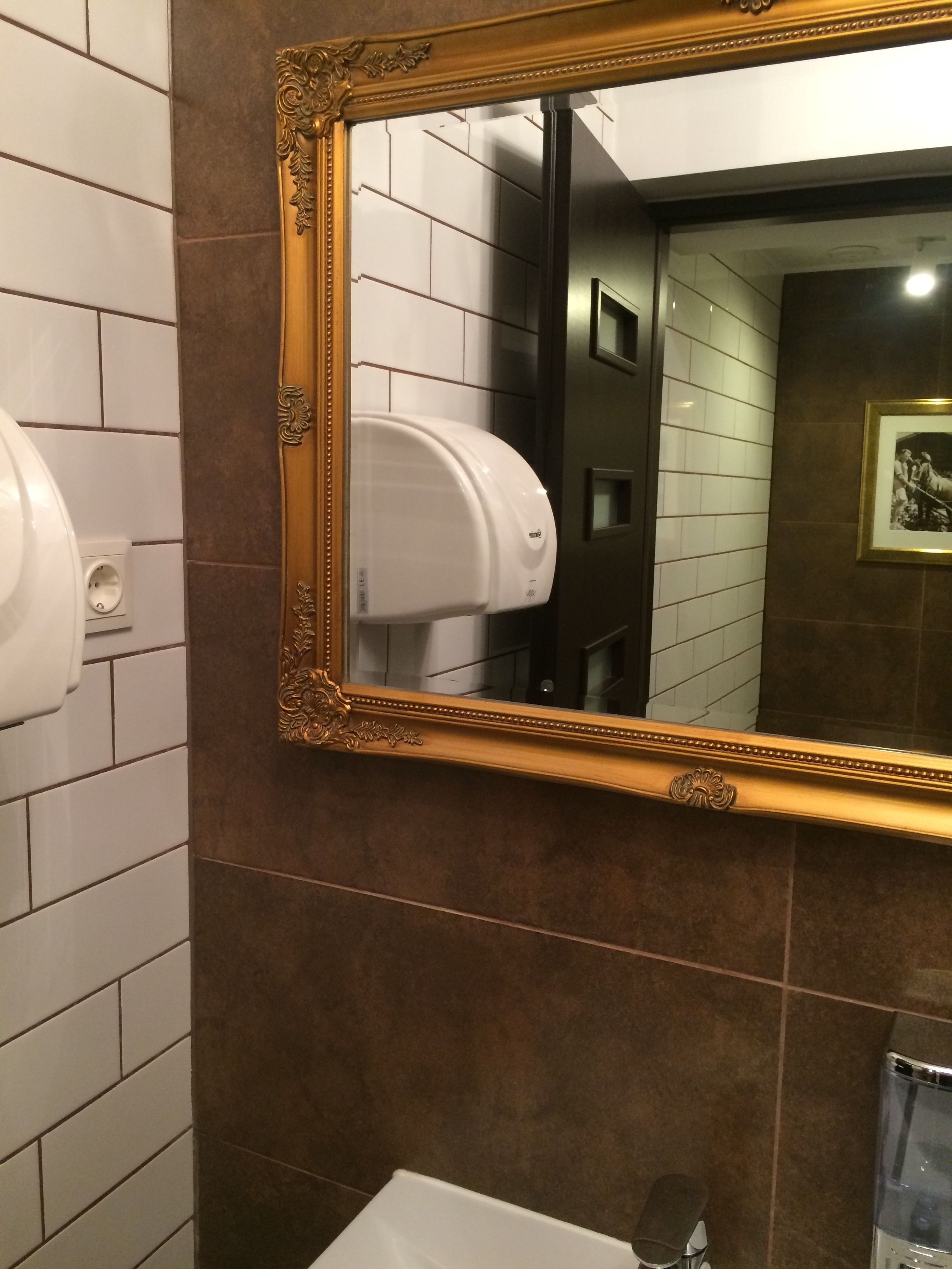 A bathroom with a large ornate mirror, a hand dryer, and a framed picture on the wall.