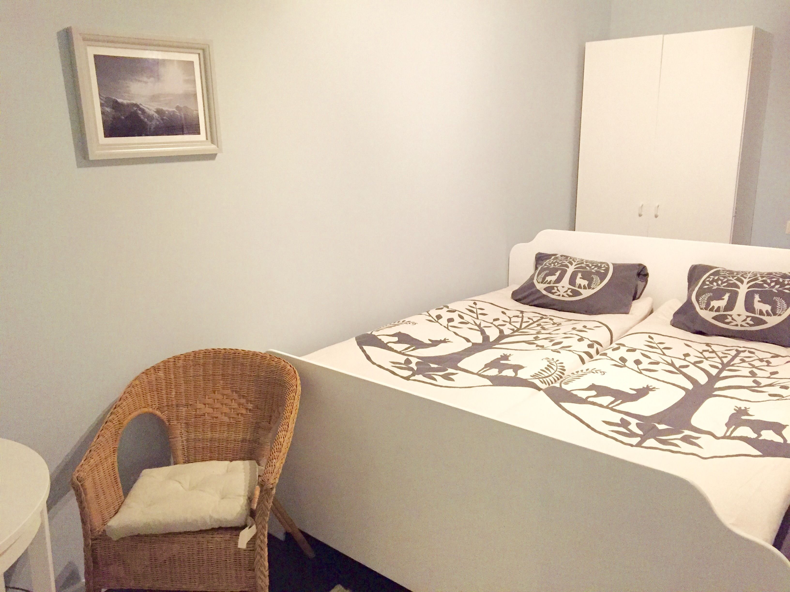 Un dormitorio con una cama, una silla de mimbre, un cuadro en la pared y un armario blanco.