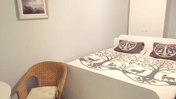 Un dormitorio con una cama, una silla de mimbre, un cuadro en la pared y un armario blanco.