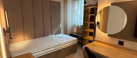 A compact bedroom with a bed, a desk, a chair, a built-in wardrobe, and a large mirror.