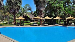 A swimming pool with lounge chairs and thatched umbrellas, surrounded by palm trees and a building in the background. - Arumeru River Lodge (Arusha)