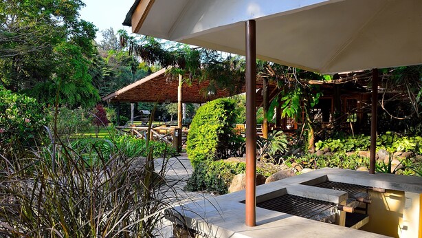 An outdoor patio area with a grill, surrounded by lush greenery and a covered seating area. - Arumeru River Lodge (Arusha)