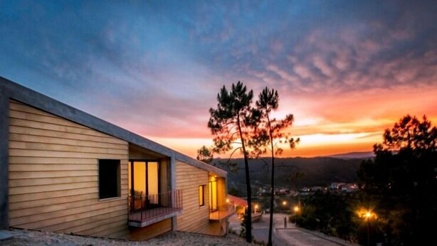 A modern house with a wooden exterior, a balcony, and a view of the surrounding landscape during sunset.