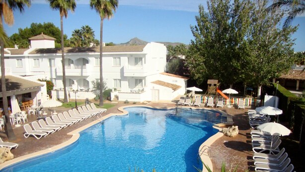 A swimming pool with lounge chairs and umbrellas, surrounded by palm trees and a building with a tiled roof. - A10 Solecito Apartamentos (Alcúdia)