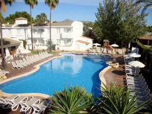 A swimming pool with lounge chairs and umbrellas, surrounded by palm trees and a building with a tiled roof. - A10 Solecito Apartamentos (Alcúdia)