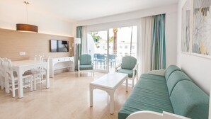 A modern living room with a sofa, a coffee table, a dining table with chairs, a TV, and a balcony with a view of buildings and palm trees. - A10 Solecito Apartamentos (Alcúdia)