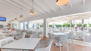 An outdoor dining area with white tables and chairs, large umbrellas, and a TV screen on the wall. - A10 Solecito Apartamentos (Alcúdia)