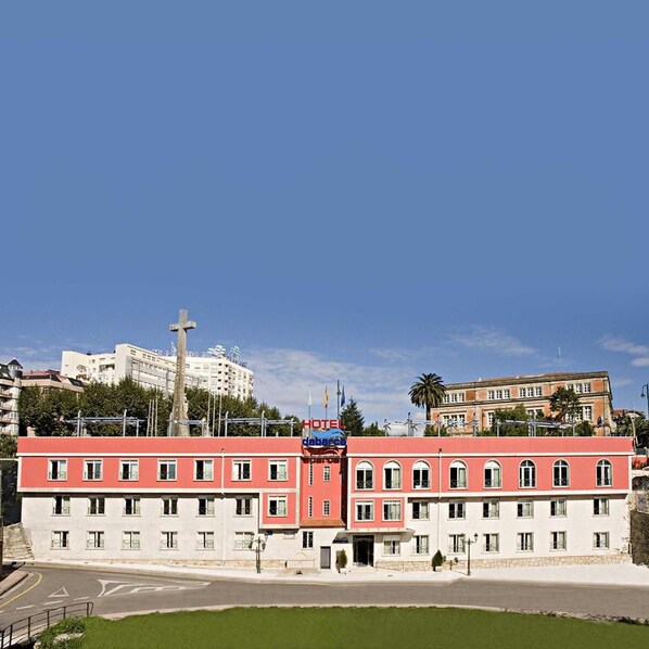 Un hotel con fachada roja y blanca, múltiples ventanas y una estructura en forma de cruz al fondo.