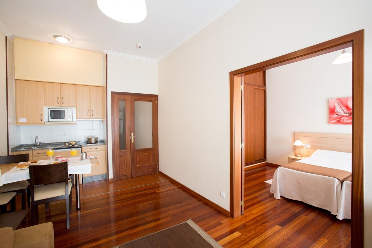A compact hotel room with a bed, bedside table, and a view of the kitchen area.