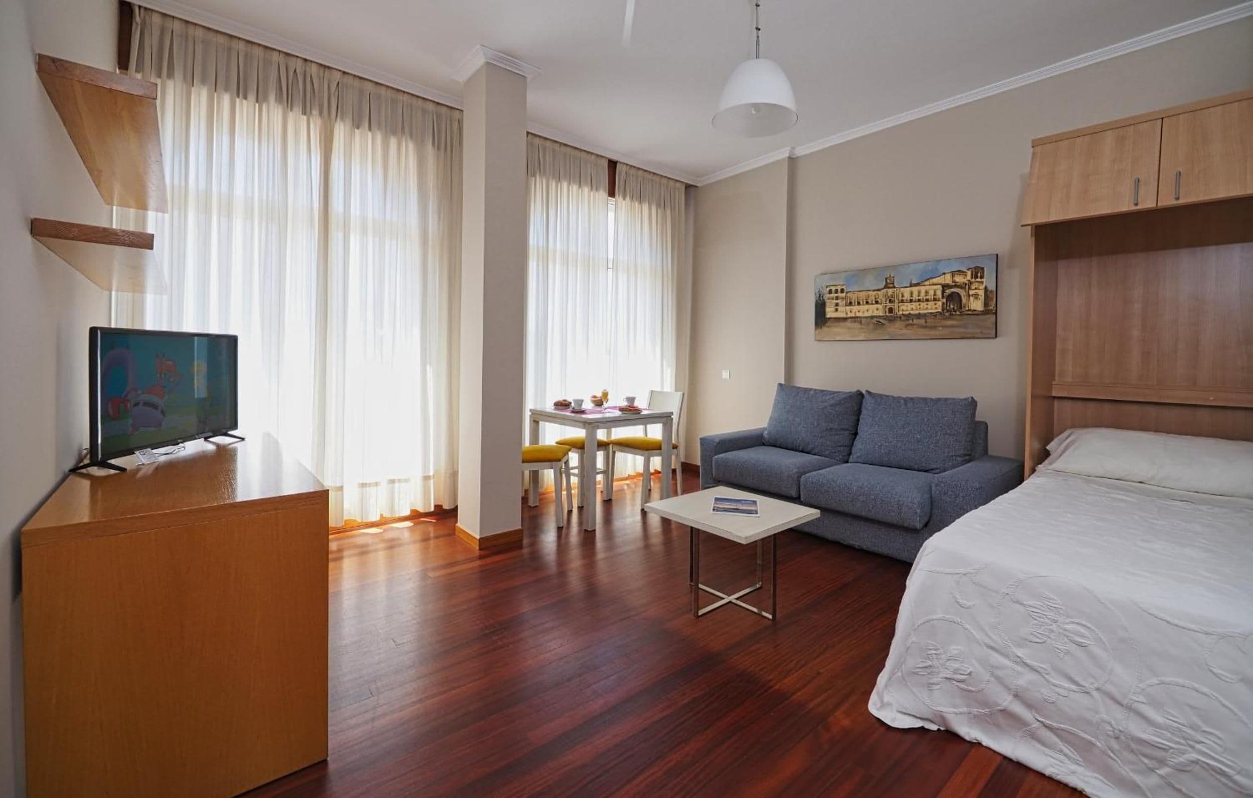 Una habitación de hotel con cama, sofá, televisión, escritorio con sillas y un cuadro en la pared.