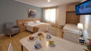 Habitación de hotel con cama, una silla, una mesa con desayuno, televisor y ventana con cortinas.