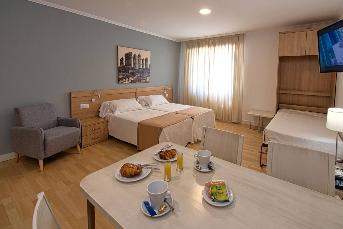 Une chambre d’hôtel avec un lit, une chaise, une table avec le petit-déjeuner, une télévision et une fenêtre avec des rideaux.