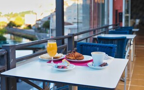 Una mesa con alimentos para el desayuno, incluyendo cruasanes, un vaso de zumo de naranja y una taza de café, frente a un balcón con vistas a edificios y una masa de agua.