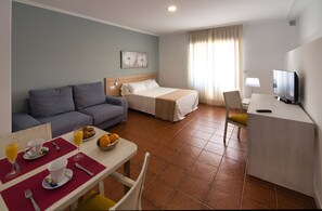 Habitación de hotel con cama, sofá, mesa de desayuno, televisión y ventana con cortinas.