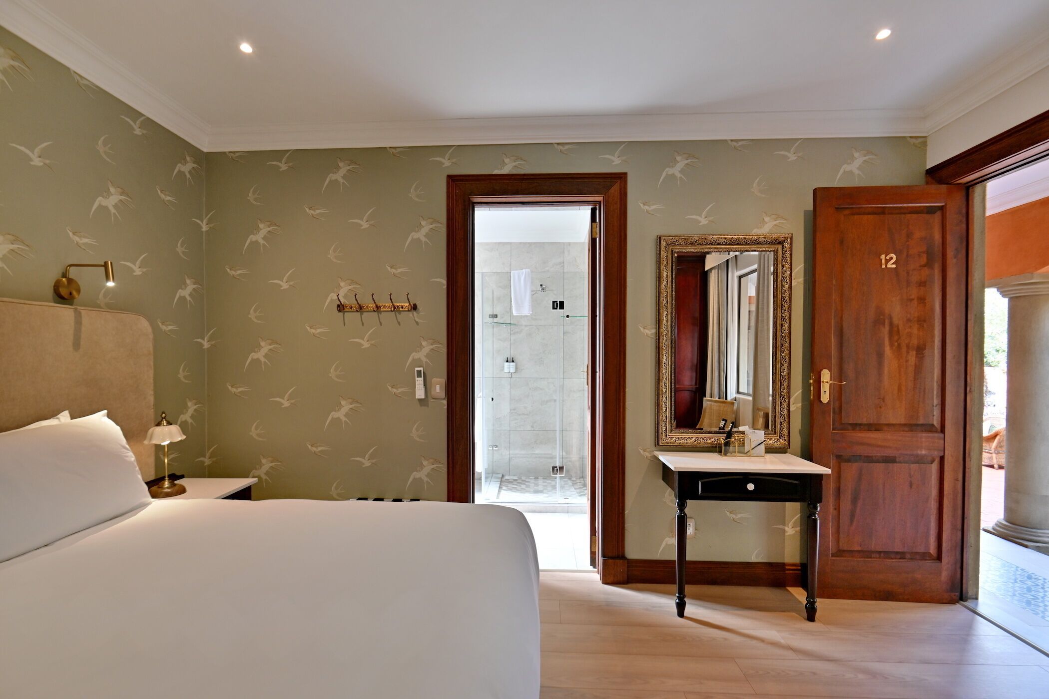 A hotel room with a bed, bedside table, lamp, mirror, and a door leading to a bathroom.