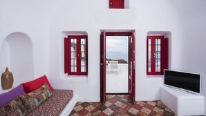 A room with a white wall, red doors, a sofa with patterned cushions, a flat-screen TV, and a view of the sea through an open door. - The Architect's Cave House (Santorini)