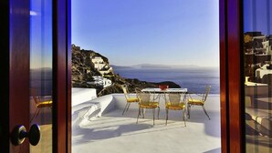 A terrace with a table and chairs overlooking a coastal landscape with white buildings and the sea. - The Architect's Cave House (Santorini)