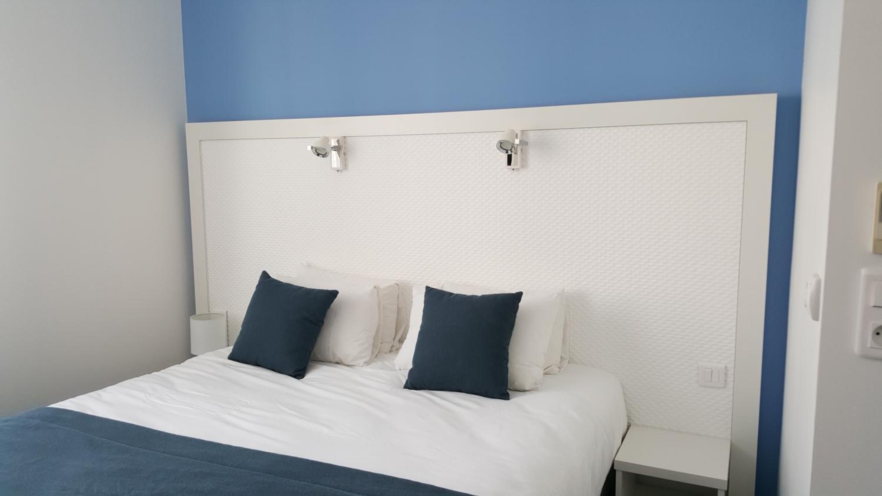 A bed with white bedding and two dark pillows, a white headboard, and a blue accent wall.