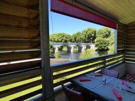 A dining area with tables set for two, overlooking a river with a stone bridge and lush greenery.